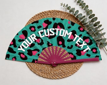 Puede incluir: Abanico turquesa con estampado de leopardo en negro y rosa. El texto blanco "YOUR CUSTOM TEXT" está impreso en la tela. El abanico abierto descansa sobre una estera tejida, con vegetación verde en el fondo.