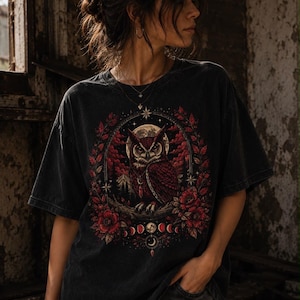 Może przedstawiać: Czarny t-shirt z krótkim rękawem z nadrukiem sowy siedzącej na gałęzi. Wzór zawiera pełnię księżyca, czerwone akcenty kwiatowe oraz fazy księżyca ułożone w okrągłej ramie z motywami gwiazd.