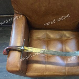 Puede incluir: Una espada curva con empuñadura de madera y guarda plateada. La hoja tiene grabados decorativos dorados. La espada descansa sobre un sillón de cuero marrón. El texto "sword crafted" es visible.