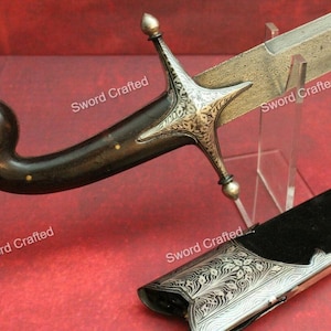 Può includere: Una spada antica con un'impugnatura curva in legno scuro ornata da borchie in ottone. L'elsa color argento presenta un intricato disegno a stella. La lama è incisa con un motivo e il fodero nero ha un'estremità decorativa argentata.