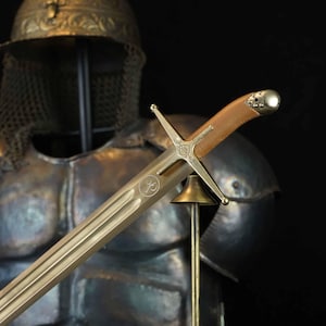Puede incluir: Espada con empuñadura de cuero marrón claro y guarda dorada. La hoja tiene un acabado dorado claro con detalles grabados. La espada se exhibe frente a una armadura.