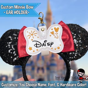 May include: Custom Minnie Mouse bow ear holder with a white bow featuring the name "Daisy" embroidered in gold thread. The bow also has gold embroidery of fireworks and balloons. The ear holder is black with silver sequins.