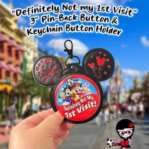 May include: Black Mickey Mouse ear-shaped keychain button holder with a 7.6 cm button. The button features Disney characters and the text "Definitely Not My 1st Visit!". The ears have embroidered designs. The background is a blurred theme park.