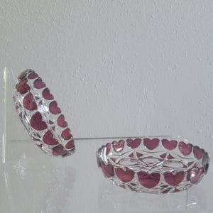 May include: Two clear glass bowls featuring a repeating pattern of deep magenta hearts around the outer rim. One bowl is positioned upright on a flat surface, while the other is tilted vertically on a clear stand to display the heart design.