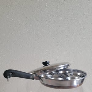 May include: A silver-toned metal pan with seven circular indentations for cooking, featuring a long black handle with a metal hanging loop and a matching silver-toned lid with a small black knob handle positioned slightly open on top.