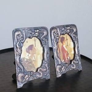 Puede incluir: Dos marcos de fotos rectangulares hechos de material metálico oscuro con un motivo de dragón en relieve que rodea una abertura ovalada. Cada marco contiene una fotografía de una persona, el izquierdo muestra a una persona de perfil y el derecho a una persona con camisa roja.