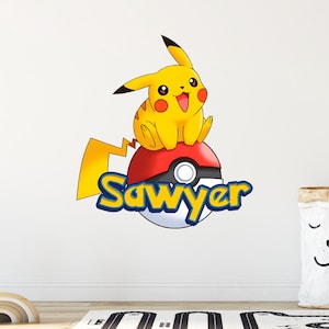 Puede incluir: Un adhesivo de pared que muestra al personaje amarillo Pikachu sentado sobre una Poké Ball roja y blanca. El nombre Sawyer está escrito en letras azules y amarillas en el centro de la bola. El diseño incluye una cola de rayo amarilla.