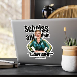 May include: A sticker on a laptop features a woman in a teal and yellow track jacket with cat-shaped designs. The sticker has the German text "Scheiss auf dein" above the woman and "KOMPLIMENT" below, with the hashtag #stopcatcalling.