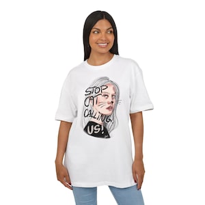 May include: White t-shirt featuring a graphic of a woman with gray hair and the text "STOP CAT CALLING US!". The illustration includes cat whiskers and a black top. The shirt is worn by a person in blue jeans.