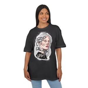 May include: Black t-shirt featuring a graphic of a woman with white hair and cat-like facial markings. The text "STOP CAT CALLING US!" is printed above the image. The shirt is a casual, oversized fit.