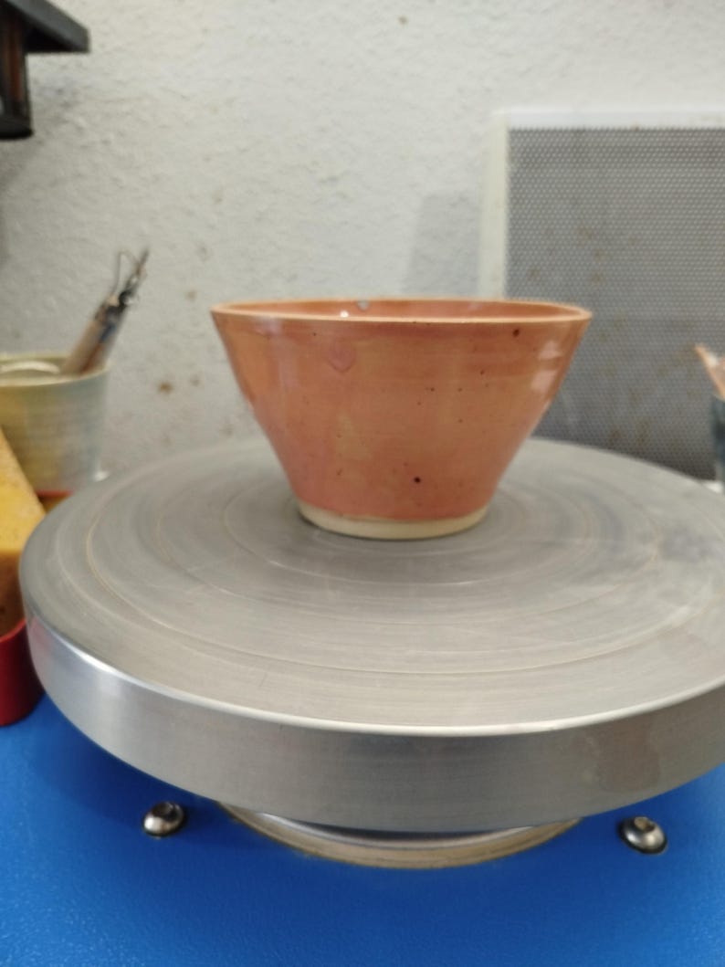 Puede incluir: Cuenco de cer&aacute;mica hecho a mano con exterior color melocot&oacute;n e interior blanco. El cuenco tiene una parte superior ancha y una forma ligeramente c&oacute;nica. Se encuentra sobre un torno de alfarero plateado.