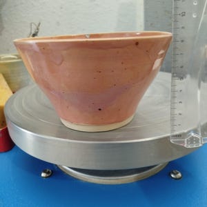 Puede incluir: Un cuenco de cer&aacute;mica melocot&oacute;n hecho a mano con una forma ensanchada. El cuenco tiene una textura moteada y una base blanca. Una regla transparente est&aacute; colocada junto al cuenco. El cuenco est&aacute; sobre una superficie met&aacute;lica plateada.