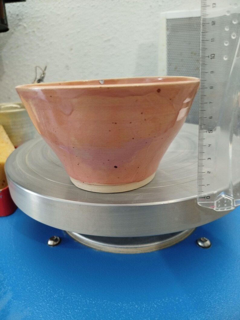 Puede incluir: Un cuenco de cer&aacute;mica melocot&oacute;n hecho a mano con una forma ensanchada. El cuenco tiene una textura moteada y una base blanca. Una regla transparente est&aacute; colocada junto al cuenco. El cuenco est&aacute; sobre una superficie met&aacute;lica plateada.