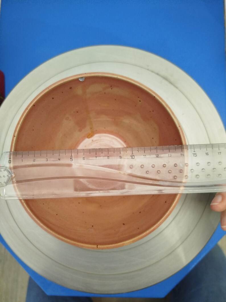 Puede incluir: Un cuenco de cer&aacute;mica hecho a mano con interior marr&oacute;n rojizo y borde marr&oacute;n claro. Una regla transparente se coloca sobre el cuenco, midiendo aproximadamente 15 cm de di&aacute;metro. El cuenco est&aacute; sobre una superficie gris y azul.