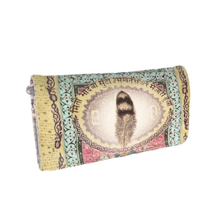 May include: A rectangular wallet featuring a central oval graphic of a brown feather surrounded by Devanagari script. The wallet has a light blue and yellow patterned border with floral motifs and a pink section at the bottom edge.