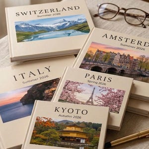 May include: A collection of travel journals with scenic cover photos of Switzerland, Amsterdam, Italy, Paris, and Kyoto. Each journal is labeled with the destination and season, such as "Summer 2026" or "Autumn 2026". A gold pen and eyeglasses are also present.