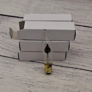May include: A stack of five white boxes with a single clear glass candle bulb with a black flame design. The bulb has a standard screw-in base.