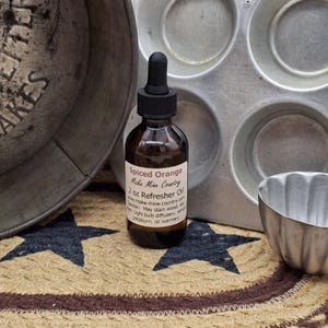 May include: A brown glass bottle of "Spiced Orange" refresher oil with a black dropper cap. The label reads "Make Mine Country" and "2 oz Refresher Oil." The bottle is next to a metal muffin tin and a small metal bowl.