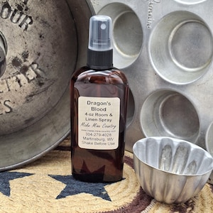 May include: A brown glass bottle of "Dragon's Blood" room and linen spray with a black spray nozzle. The label includes the text "Make Mine Country." The bottle is next to a metal muffin tin and a small metal mold.