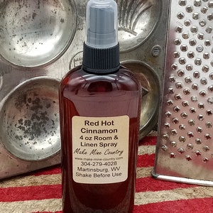 May include: A brown glass bottle with a black spray nozzle and a white label. The label reads "Red Hot Cinnamon 4 oz Room & Linen Spray Make Mine Country www.make-mine-country.com 304-279-4028 Martinsburg, WV Shake Before Use".