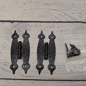 May include: Two black metal hinges with a hammered finish and pointed ends. The hinges are attached to a white wooden surface. A bag of screws is included.