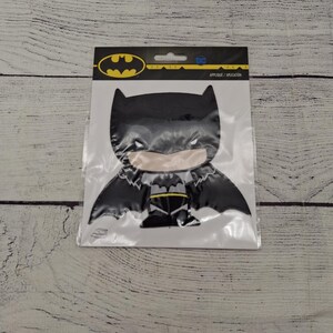 May include: A black Batman applique on a white background, packaged in a clear plastic sleeve. The applique features the Batman logo and the DC logo. The applique is a cartoon-style design.