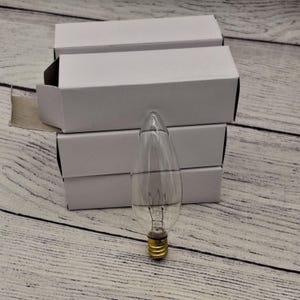 May include: Four white boxes stacked on top of each other with a clear glass light bulb in front of them. The light bulb has a traditional filament and a standard screw base.