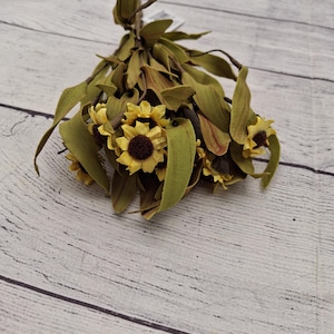 May include: A decorative bundle of artificial flowers featuring small yellow sunflowers with dark brown centers, interspersed with long, pointed olive green leaves. The stems are gathered at the top, creating a compact, rounded arrangement of botanical elements.