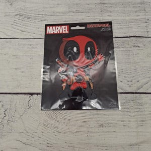 May include: A Deadpool applique patch on a black background. The patch features a cartoon version of Deadpool in red and black, with the Marvel logo in the upper left corner. The words "DEADPOOL" and "APPLIQUE APLICACION" are printed on the top right.