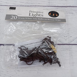 May include: A package of 20 warm white tiny bulbs on brown wire primitive lights. The package is labeled "Primitive Lights" and "20 Warm White Tiny Bulbs on Brown Wire".