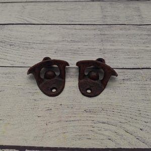 May include: Two cast iron bottle openers with a rustic, reddish-brown finish. Each opener has a unique design with a curved top, a central ball, and a small hole at the bottom for mounting. The openers are designed to open bottles.