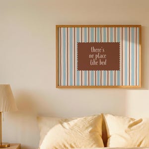 Pode incluir: Uma impressão de arte emoldurada com uma moldura de madeira marrom. A obra de arte apresenta um fundo listrado em azul e marrom, com um retângulo marrom no centro que diz "there's no place like bed."