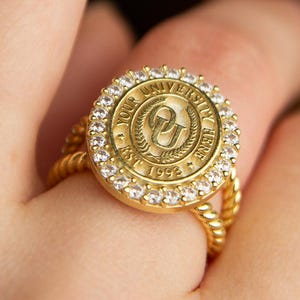 May include: A gold-colored ring with a university seal design. The ring features a round, detailed emblem surrounded by clear gemstones. The band has a twisted rope design. The ring is worn on a finger.