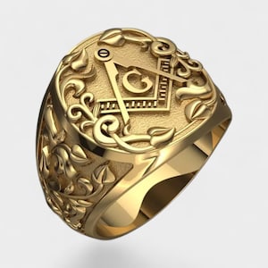 Può includere: Un anello color oro con un emblema massonico. L'anello presenta un design a squadra e compasso con una 'G' al centro, circondato da dettagli floreali ornati. L'anello è mostrato su sfondo bianco.