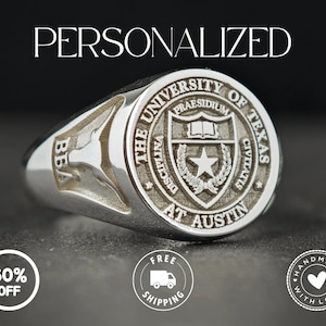 May include: A silver ring with the text "PERSONALIZED" at the top. The ring features the University of Texas at Austin seal and the letters "BBA". The ring is on a dark surface.