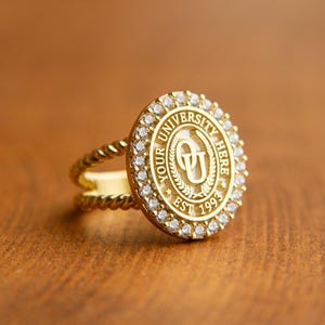 May include: A gold ring with a university emblem, encircled by small clear stones. The ring features a twisted band and a round face with the words "YOUR UNIVERSITY HERE EST 1993". The ring is set on a wooden surface.