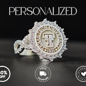 May include: A silver ring featuring the Texas Tech University logo. The ring has a round face with the university name and logo, surrounded by clear gemstones. The band is also adorned with gemstones. The word "PERSONALIZED" is at the top.