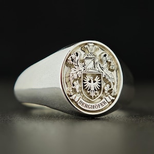 May include: A silver signet ring with a detailed crest engraving. The crest features a shield with an eagle, surrounded by ornate details and the name "BERGHÖFER" in a circular banner. The ring has a smooth, polished finish.