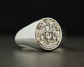 Può includere: Un anello con sigillo d'argento con un'incisione dettagliata di uno stemma. Lo stemma presenta uno scudo con un'aquila, circondato da dettagli ornati e il nome "BERGHÖFER" in un banner circolare. L'anello ha una finitura liscia e lucida.