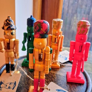 May include: A selection of hand-painted wooden nutcrackers in colours such as gold, green, and red. Each figure has a unique hat design and stands on a small wooden base. Some have tags attached.
