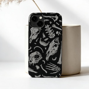 May include: Black phone case featuring a repeating pattern of gray animal bones and skulls. The case is made of a hard, protective material and is designed to fit a smartphone. The design includes various skeletal elements.