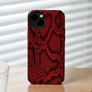 Pode incluir: Uma capa de telefone vermelha com padrão de pele de cobra. A capa está em pé sobre uma superfície de madeira. A capa do telefone tem um recorte para a câmera e uma lente preta. Um relógio com pulseira de couro marrom está em primeiro plano.