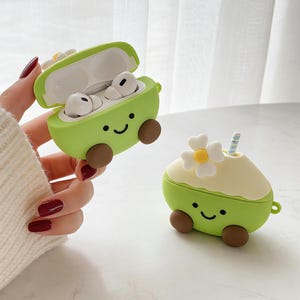 Peut inclure: Étui AirPod en silicone vert clair en forme d'avocat, avec des roues marron et un visage souriant. L'étui est ouvert, révélant des écouteurs blancs. Un autre étui en forme d'avocat avec une fleur et une paille est également visible.
