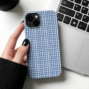 May include: A blue and white checkered phone case. The case is held near a laptop keyboard. The case has a grid pattern of white lines on a blue background. The phone case has a camera cutout.