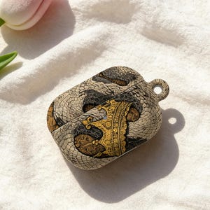 Puede incluir: Una funda decorativa para auriculares inalámbricos, con un diseño de piel de serpiente en tonos grises, marrones y beige. El diseño incluye una corona dorada. La funda tiene un pequeño lazo para sujetarla a un llavero o cordón.