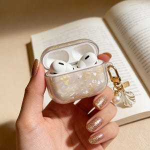 Może przedstawiać: Przezroczyste etui na AirPody z perłowym i złotym wzorem. Etui jest otwarte, ukazując białe AirPody. Złoty urok z perłą i muszlą wisi na etui. Etui jest trzymane w dłoni z błyszczącym złotym lakierem do paznokci.