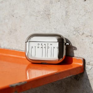 May include: A shiny, silver-colored AirPods case with the words "DEATH STRANDING" printed in black. The case is sitting on an orange shelf against a gray wall. The case has a smooth, reflective surface.