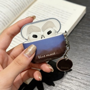 Puede incluir: Una funda para AirPods con degradado azul y marrón con el texto "blue mood." La funda está abierta, revelando unos AirPods blancos. Un pequeño dije redondo marrón cuelga.