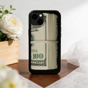 May include: Black phone case displaying a stack of US $100 bills, held together by a white rubber band. The phone case has a black frame and a camera cutout. The bills are visible through the case.