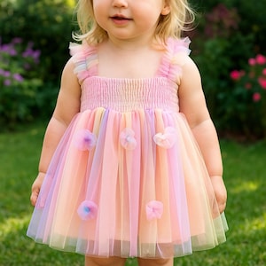 May include: A pastel rainbow-colored toddler dress with a tulle skirt and floral accents. The dress features a square neckline, ruffled straps, and a gathered bodice. The skirt has vertical stripes of pink, yellow, and blue.